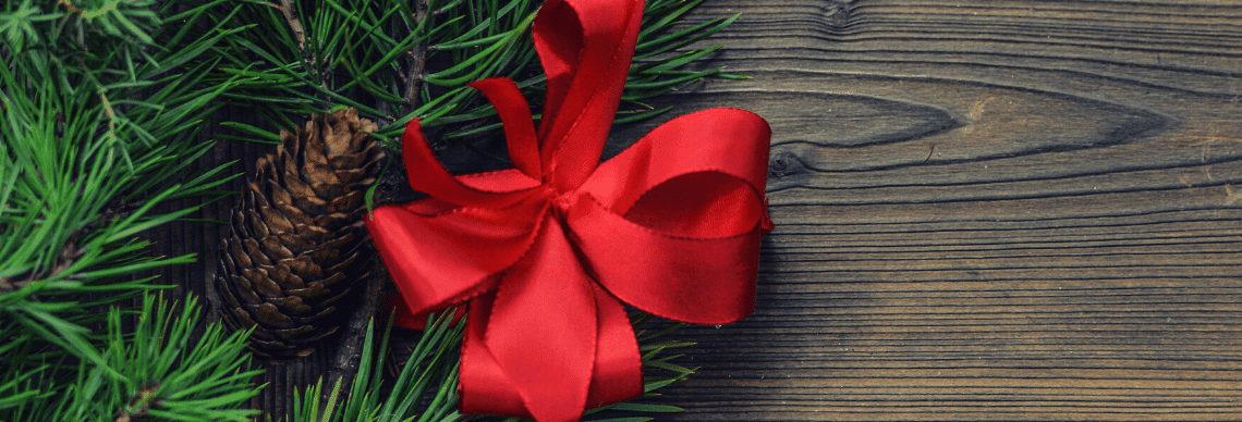 A bright red ribbon tied into a bow, resting on a bed of green pine needles. To the left of the bow, there is a pine cone nestled among the needles, adding a natural, rustic element to the composition.