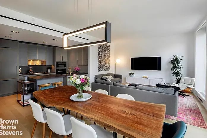 Open-plan living room with a gray sectional, long wood dining table, and modern kitchen.