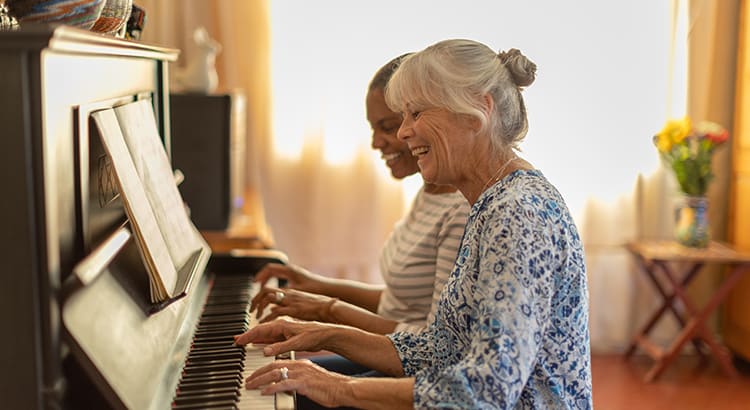  An elderly woman playing the piano, possibly teaching or playing along with a younger person. They are in a warmly lit room, suggesting a home setting.