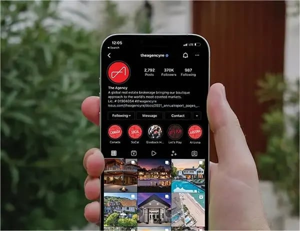  A hand holds a white smartphone, displaying an Instagram profile page for "theagencyre" against a blurred background of green foliage and a dark red wall.