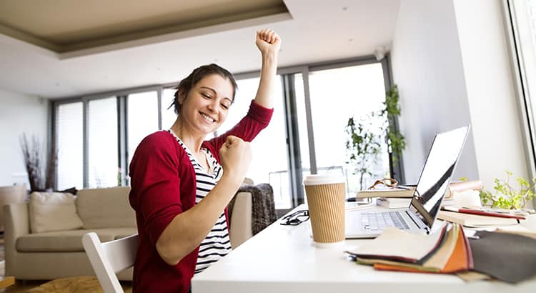 A woman in a cheerful pose, raising her fist in a gesture of success or celebration, seated at a table with a laptop and a cup, likely representing a moment of accomplishment while working from home.