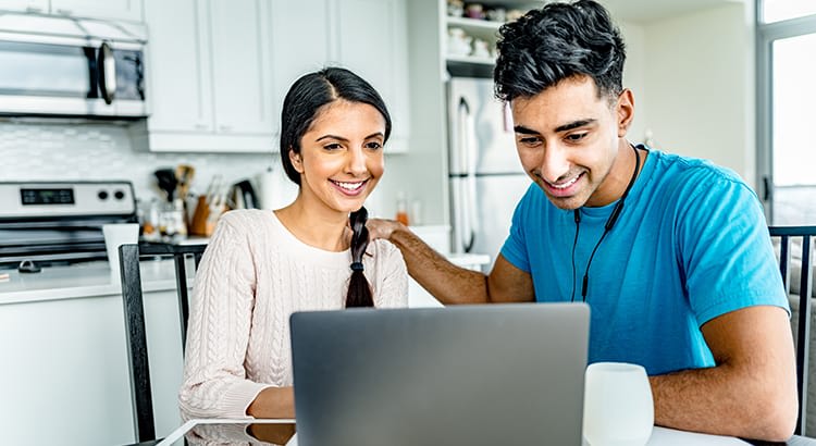 A smiling couple sits together in a kitchen or dining area, engaging with a laptop, perhaps discussing plans or working on something together.