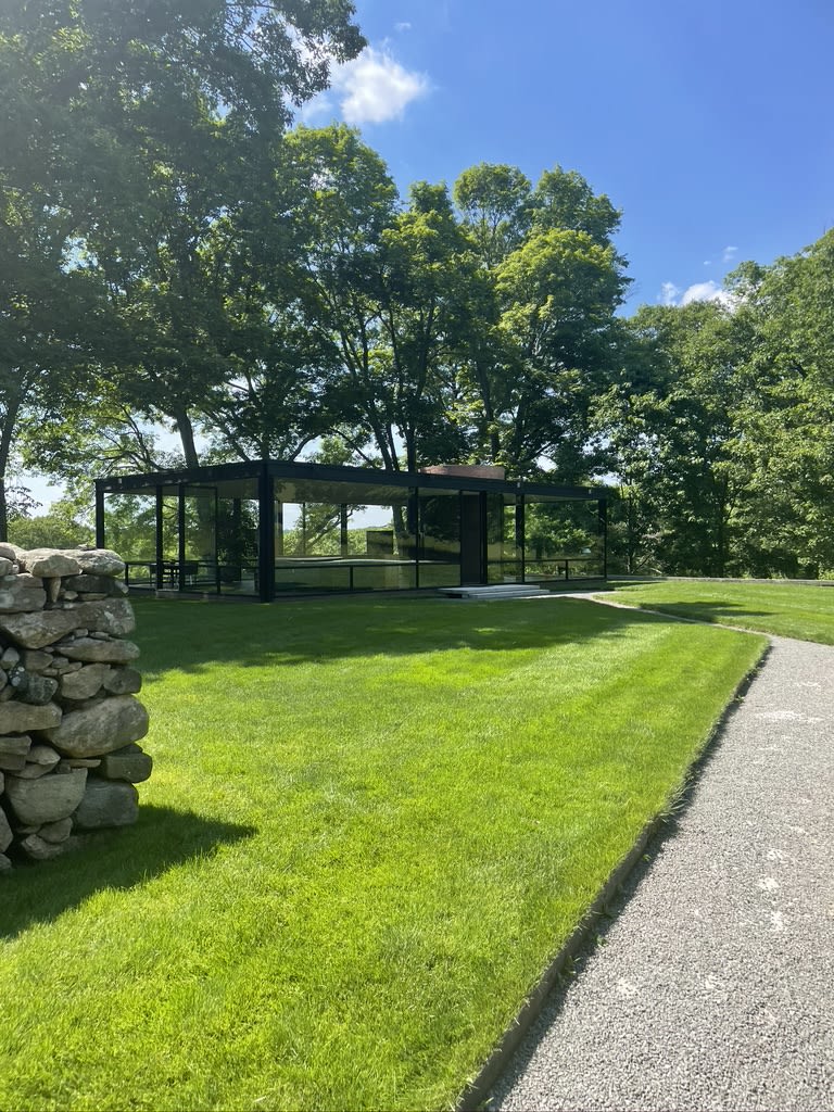 A modern glasshouse in the middle of a grassy field. The house has floor-to-ceiling windows and a black metal frame.