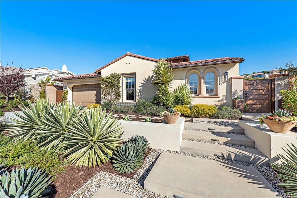 Front view of a Mediterranean-style luxury home with desert landscaping, featuring agave plants and a tiled roof.