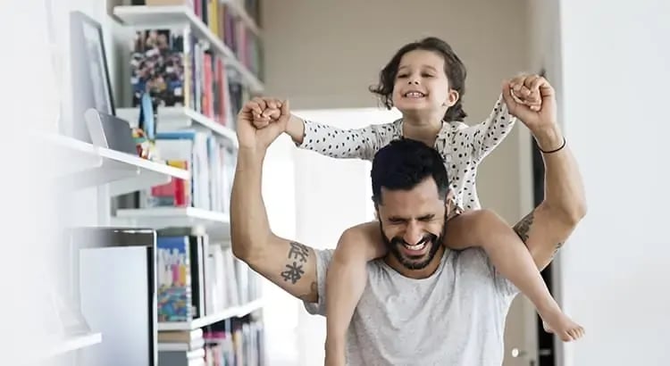 A joyful moment featuring a man with a child on his shoulders, both smiling broadly. The scene is indoors, in a well-organized room with bookshelves in the background. It conveys happiness and family bonding.