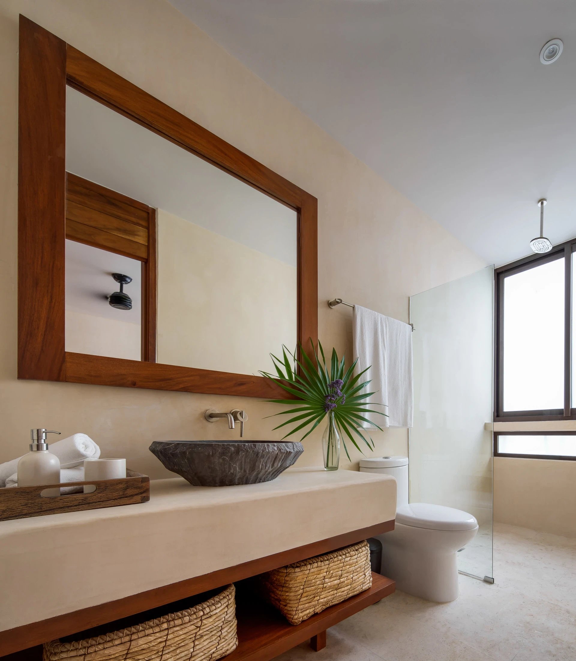 Bathroom with a wood-framed mirror, stone vessel sink, woven baskets, and glass-enclosed shower.