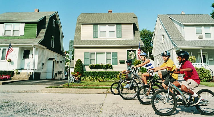 A street view of a neighborhood with children riding bicycles. The houses have a classic architectural style, with well-maintained lawns and trees.