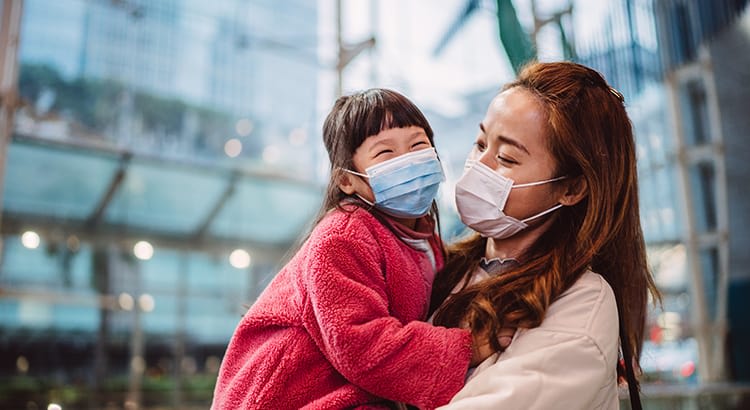 A joyful moment between a mother and her young child, both wearing face masks, likely in an urban setting. The child, dressed in a bright pink jacket, is smiling through the mask, while being held by the mother.