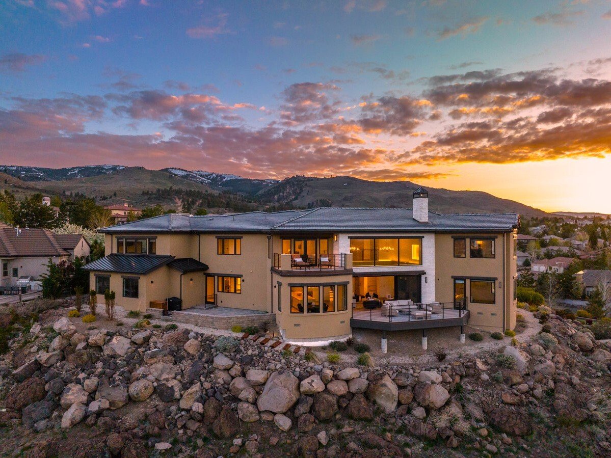 Modern two-story home with broad windows and multiple decks on a rocky hillside at sunset.