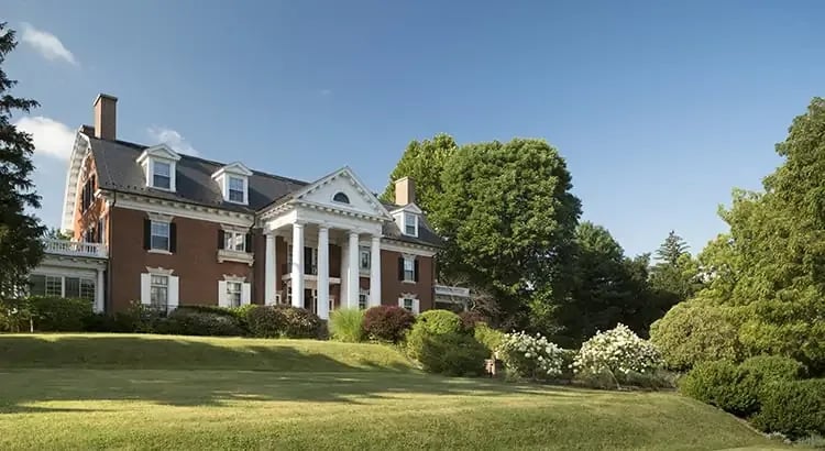 A grand estate with a large, elegant house surrounded by a vast green lawn and trees. The scene suggests luxury, affluence, or historical significance.