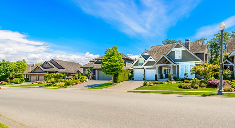 A suburban street lined with modern houses. The homes have well-manicured lawns, and the sky is clear and blue.