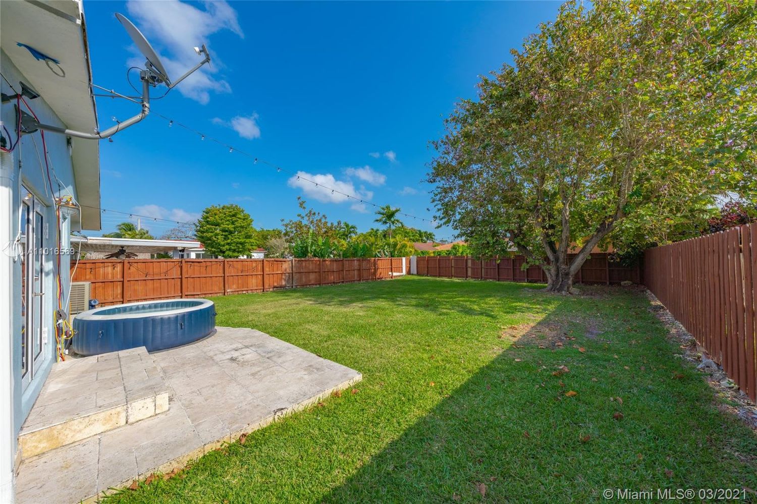 Fenced backyard with a large lawn, flowering tree, and round spa beside a stone patio.