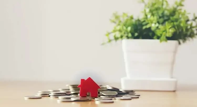  A small model house placed on a table next to stacks of coins, symbolizing real estate investment or saving for a home.