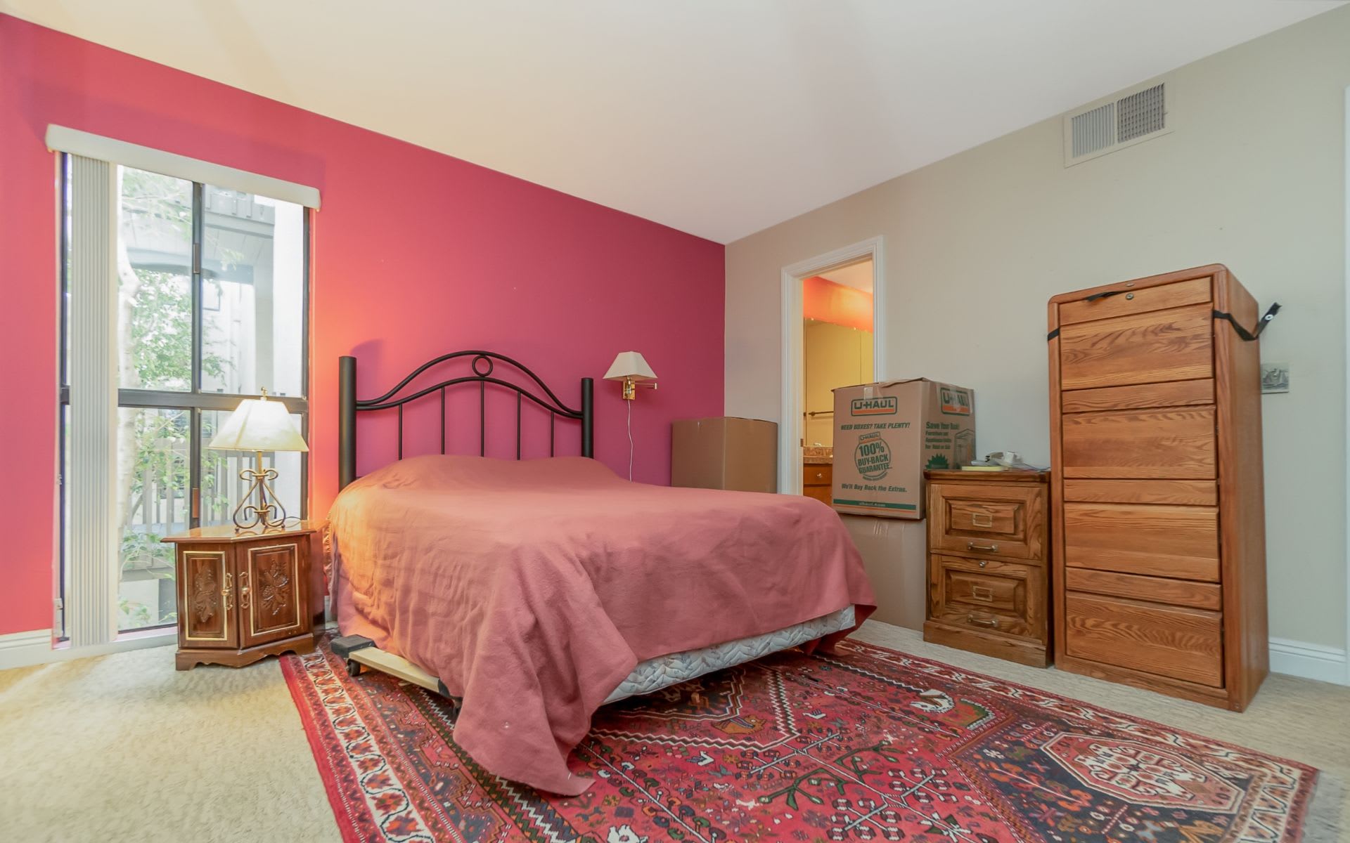 Bedroom with a pink accent wall, black metal bed, wooden dressers, and moving boxes.