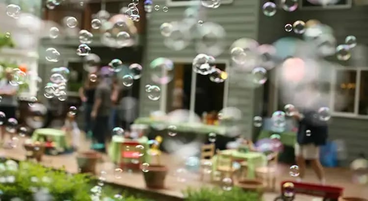 A blurred image of an outdoor market or festival with people in the background. The foreground features bubbles floating, adding a playful and whimsical element to the scene.