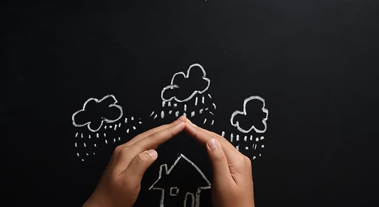A symbolic image showing hands forming a protective gesture over a small house, with illustrations of rain and clouds above, possibly symbolizing insurance or protection.