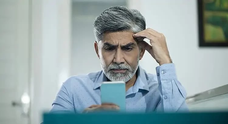 An elderly man with gray hair and a beard, looking at a smartphone with a concerned expression. He is wearing a light blue shirt, and the background is blurred, possibly an indoor setting.