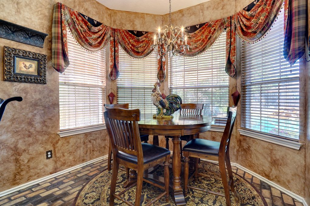 Dining nook with a round wooden table, rooster centerpiece, chandelier, and large curtained windows.