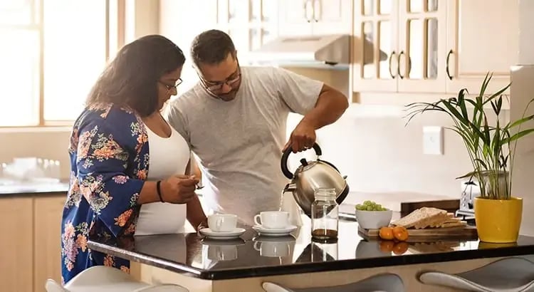 A couple cooking together in a modern kitchen. The man is pouring something from a kettle while the woman is chopping vegetables, indicating a shared and cooperative activity.