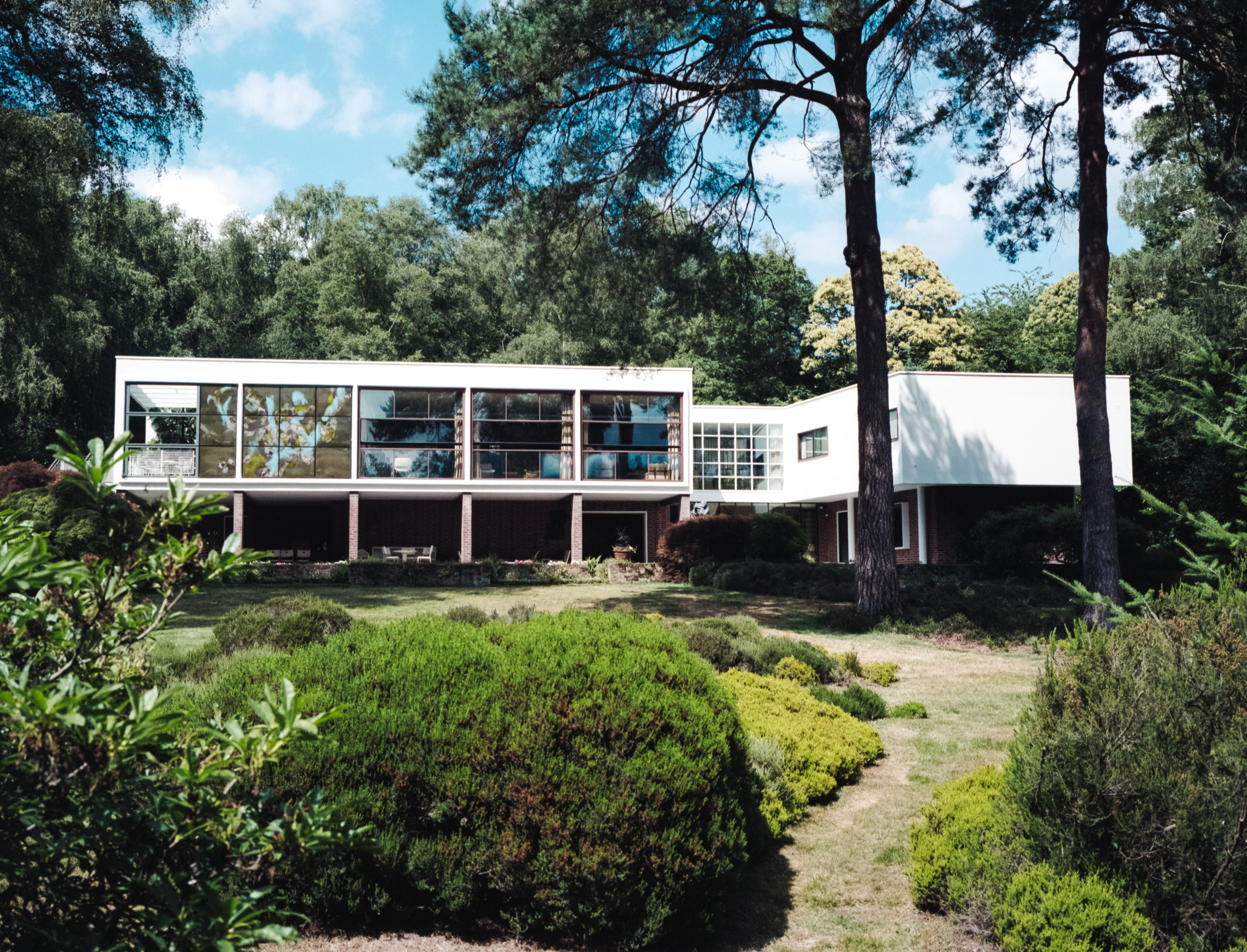 Modern white-and-brick home with broad windows, mature trees, and landscaped grounds.