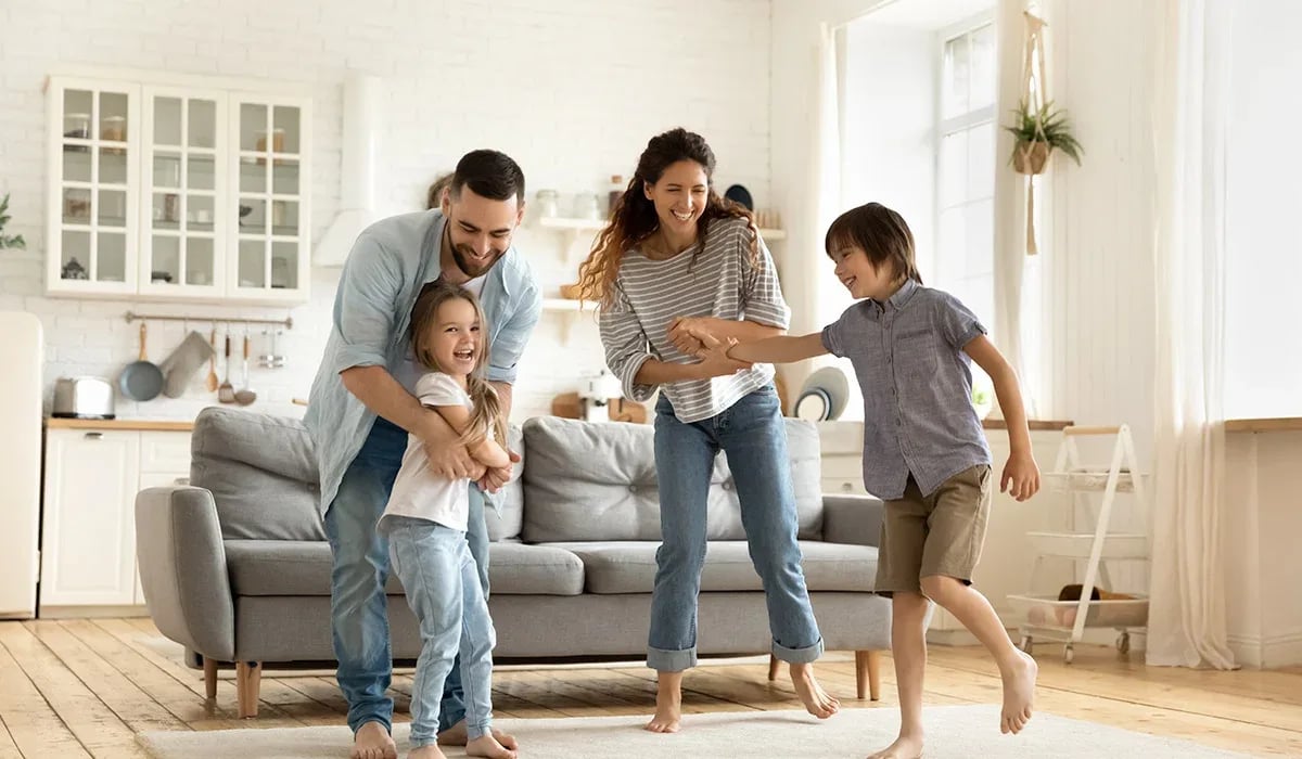 Family of four playing together beside a gray sofa in a bright living room.