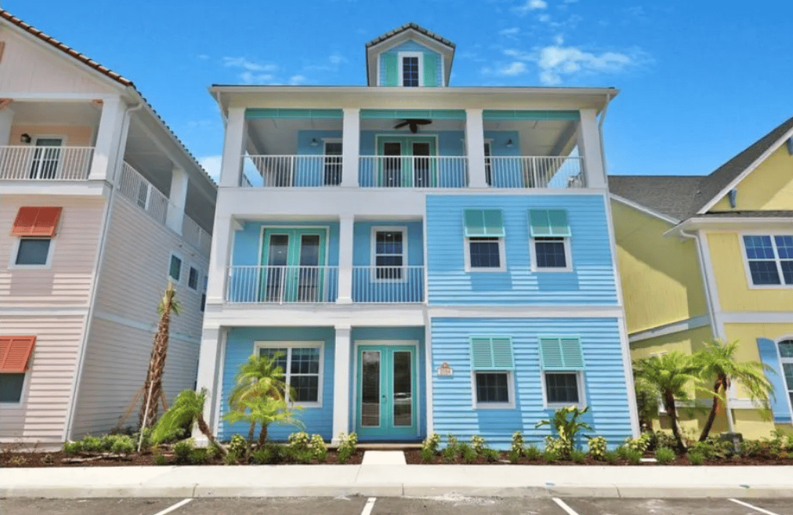 A blue and white two-story house with a balcony, a walkway, flower beds, trees, and other homes in the background.