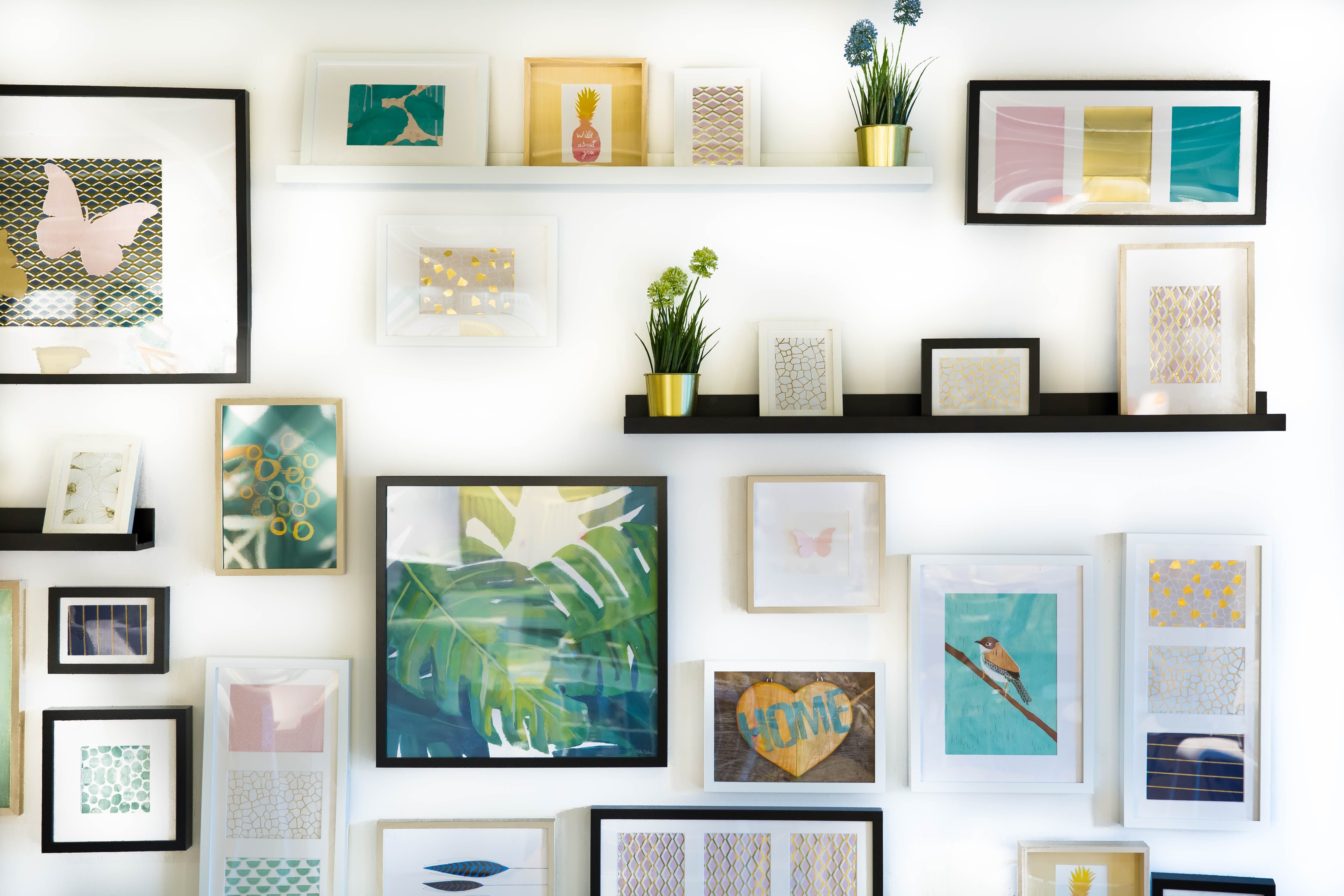 Decorated white wall with colorful framed art, floating shelves, and potted plants.
