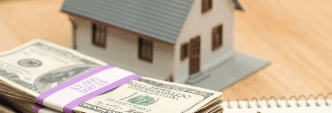 A miniature house sits on a flat surface, representing real estate or property ownership. In the foreground, there is a bundle of US dollar bills, secured with a purple strap labeled "$2,000".