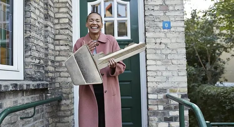 A woman standing outside, holding a large key. She is smiling and appears to be in front of a brick building, possibly symbolizing moving into a new home or real estate.