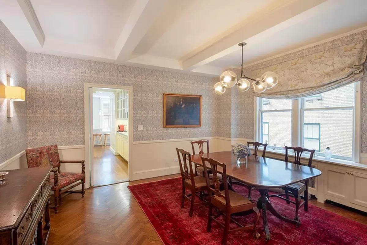 Dining room with a dark wood table on a red rug, patterned wallpaper, and large windows.