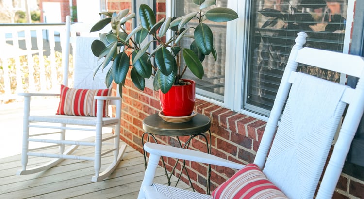 A cozy porch scene with two white wooden rocking chairs positioned near a window and a brick wall. 