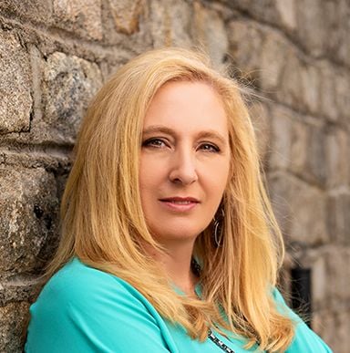 Laura Hart with long blonde hair and a turquoise top against a stone wall.