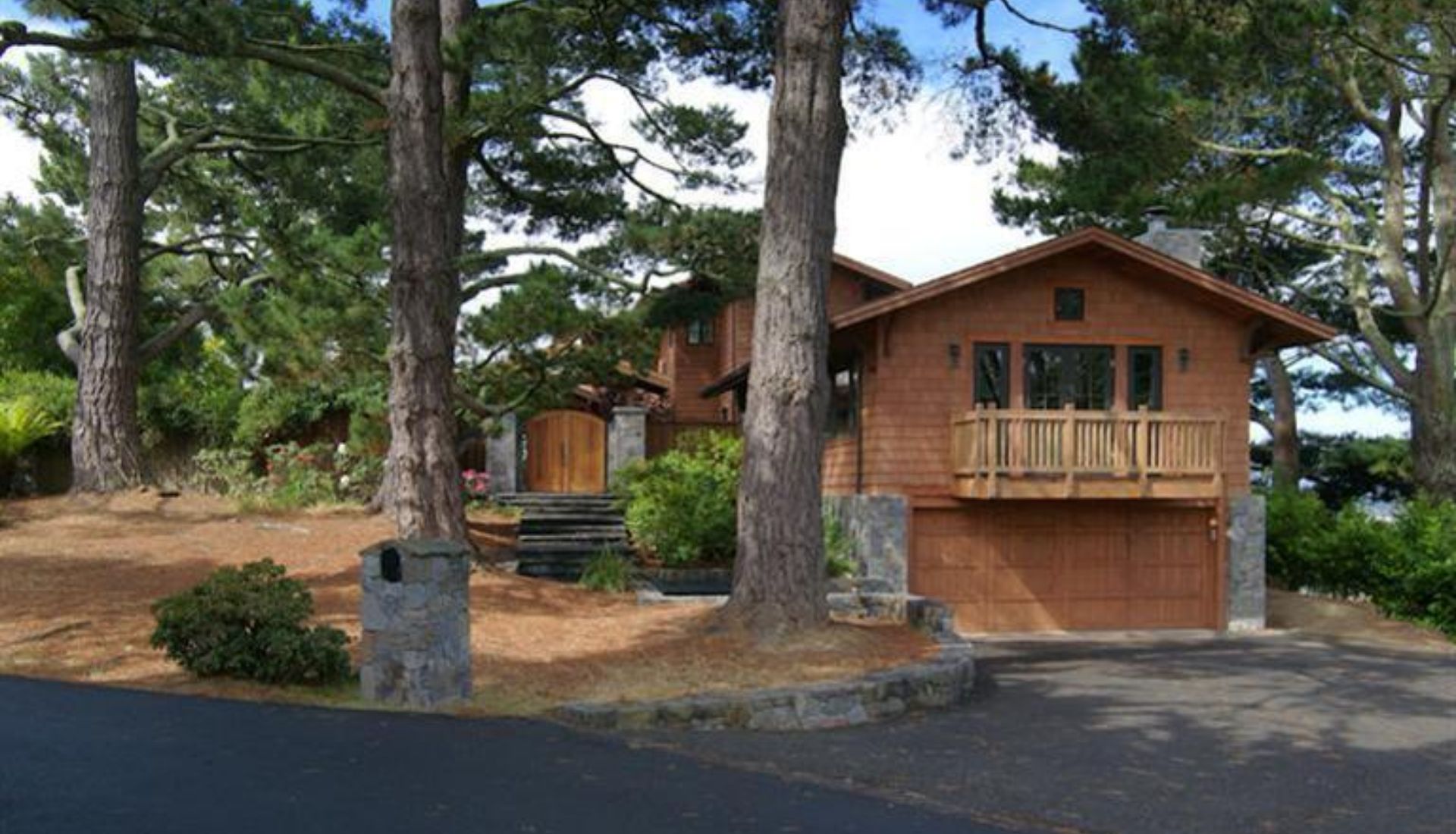 Cedar-shingle house with a wooden balcony, garage, stone entry pillars, and mature trees.