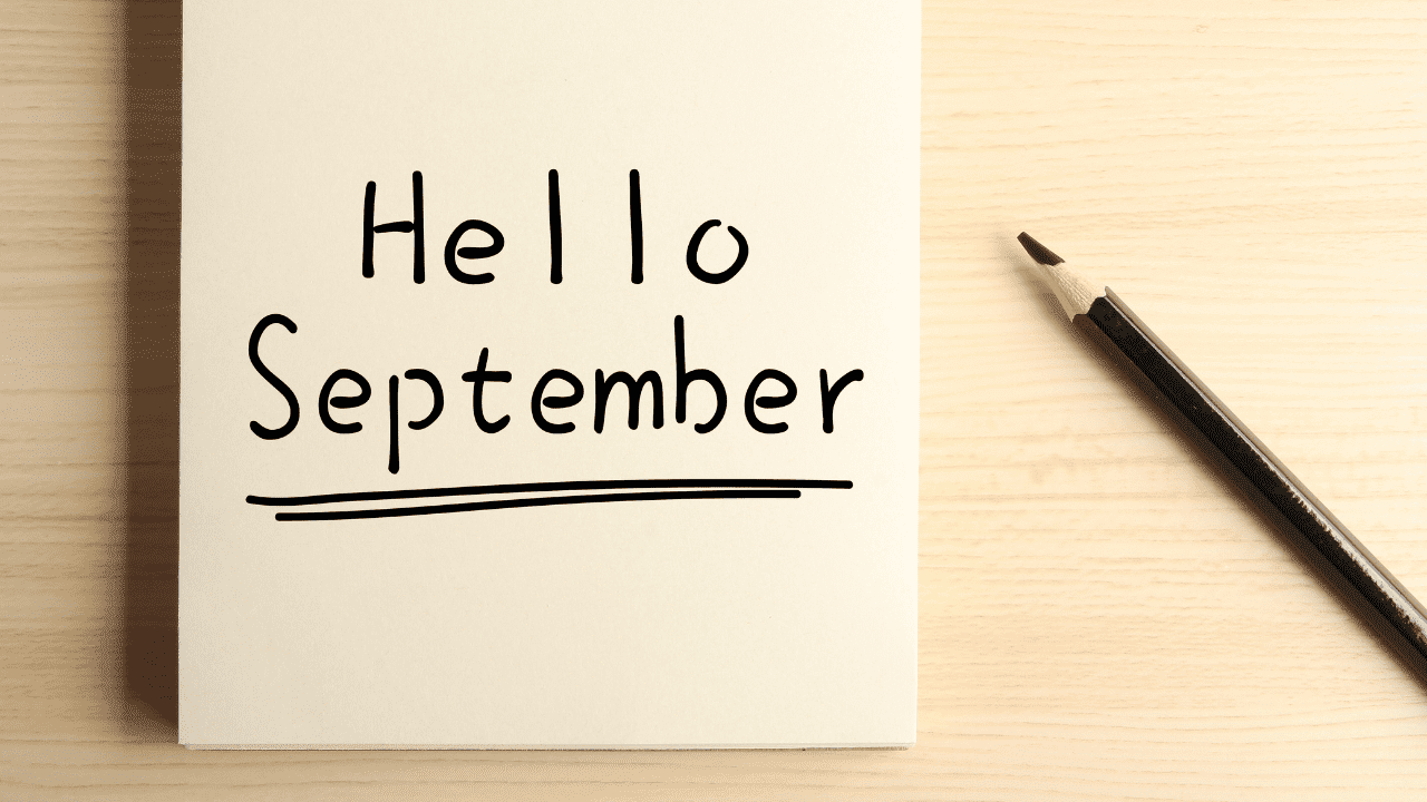 On a wooden surface, there is a piece of paper or card with the words "Hello September" written in black ink. 