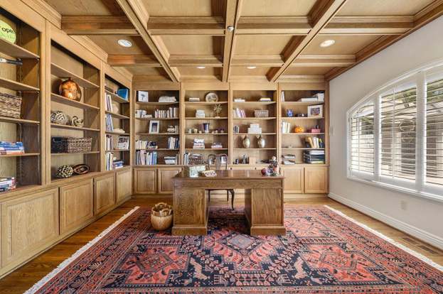 Wood-paneled home office with built-in bookshelves, coffered ceiling, central desk, and large shuttered windows.