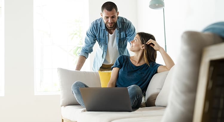 A casual and intimate moment between two people, likely a couple. One person is sitting on a couch with a laptop on their lap, while the other stands beside them, leaning forward slightly in a conversational posture. 