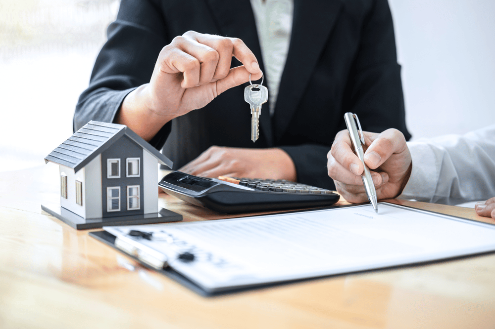 Model house, keys, calculator, and paperwork on a desk as someone writes on a document.