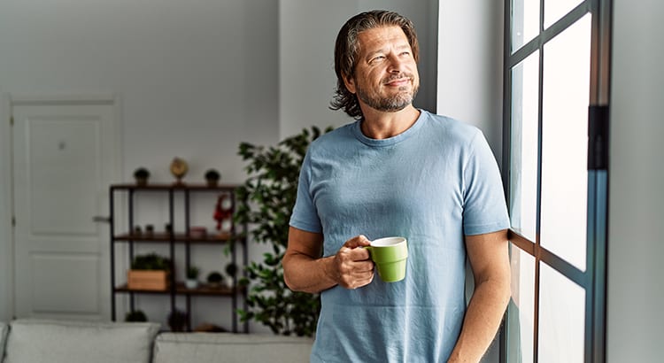 A man standing in a modern, well-lit room, holding a coffee mug. He is casually dressed and smiling, with shelves and plants visible in the background.