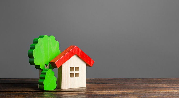 An image of a small wooden house model with a red roof next to a small green tree on a wooden surface, symbolizing real estate or homeownership concepts.