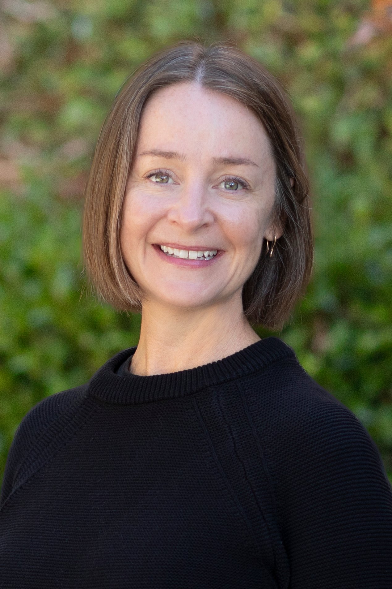 Anastasia Beckett smiling in a black sweater against a leafy green background.