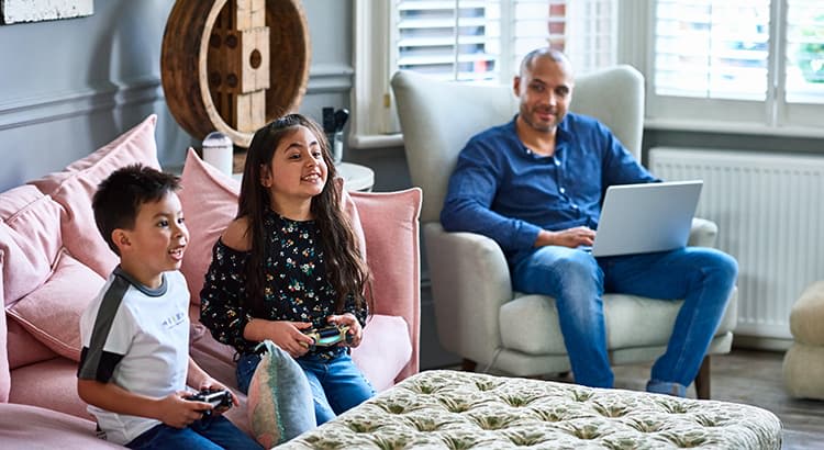 wo children, a boy and a girl, are sitting on a couch, enthusiastically playing a video game using controllers. They both appear happy and engaged in the game. Behind them, an adult man (possibly their father) is sitting on a comfortable chair, working on a laptop. The room is well-lit with natural light coming through the window blinds, and the furniture appears to be soft and inviting. The atmosphere is relaxed, combining work and play in a family setting.