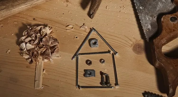 The central focus is a simple house shape constructed from nails and small metal parts. The nails form the outline of the house, with additional metal pieces inside representing windows and a door. To the left of the house shape is a small pile of wood shavings, resembling a tree with a wooden stick as its trunk.