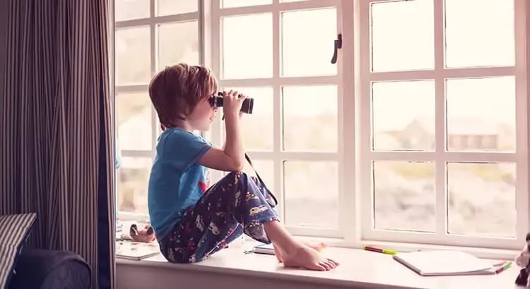 A child, likely a boy, sitting by a large window with binoculars, looking outside. The child is in pajamas, indicating it might be morning. This scene conveys curiosity and exploration.