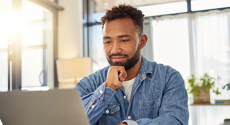 A man sitting at a desk with a thoughtful expression, working on a laptop, suggesting themes of remote work, professional development, or market analysis.
