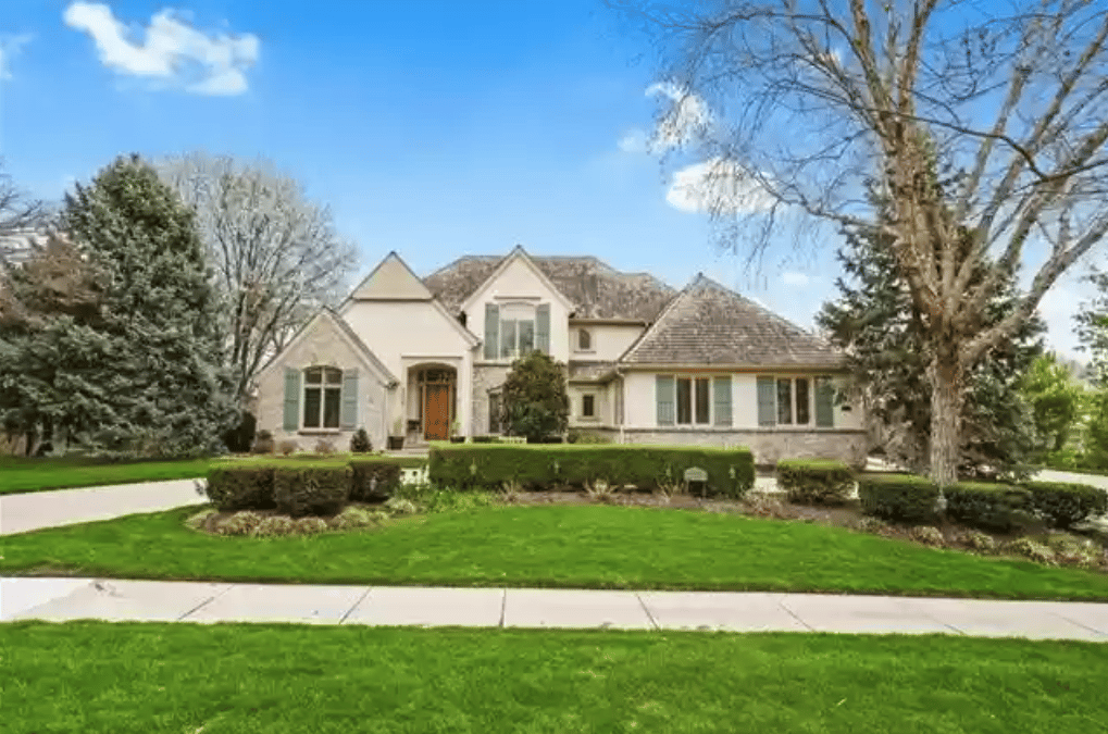 Two-story cream house with gabled rooflines, gray shutters, and a broad green front lawn.