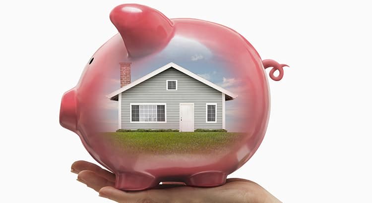 A pink piggy bank being held in the palm of a hand. Inside the transparent piggy bank, there is a house depicted, symbolizing saving for a home or homeownership. 