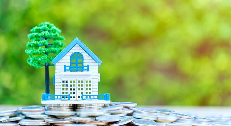 The house is white with blue accents, featuring windows, a door, and a small balcony. Next to the house is a model tree, suggesting a residential setting. The background is blurred, with greenery visible, creating a peaceful outdoor atmosphere. The scene conveys a connection between homeownership and financial investment, symbolized by the house sitting atop the stack of coins.