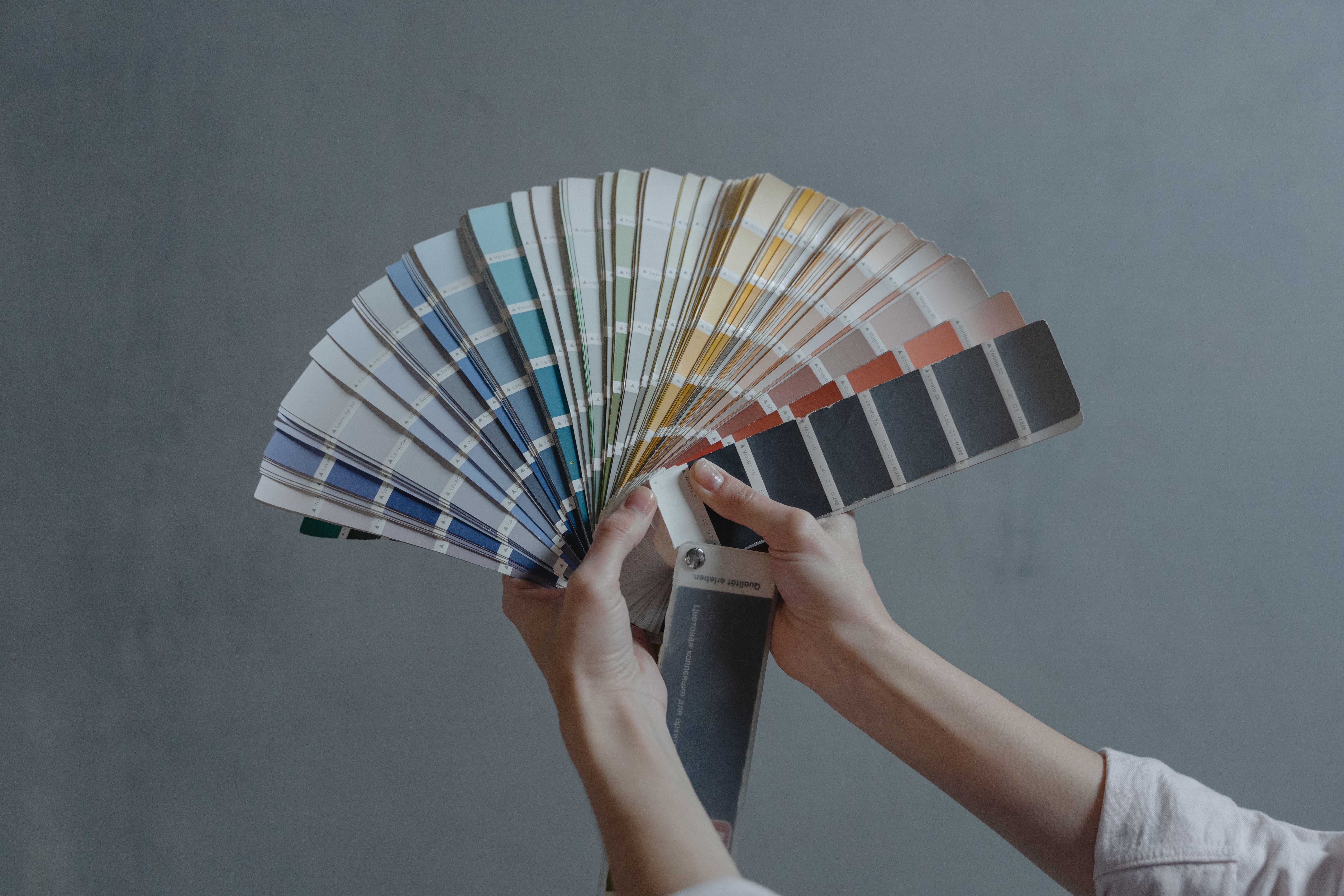 Hands fan out paint color swatches in a range of hues, including blues, warm reds, and charcoal.