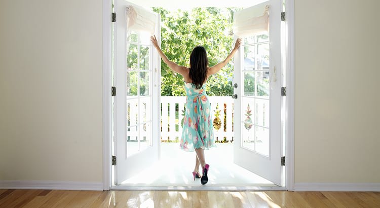 A woman standing in front of a wide set of open French doors leading to a bright and sunny outdoor space, possibly a balcony or patio. 