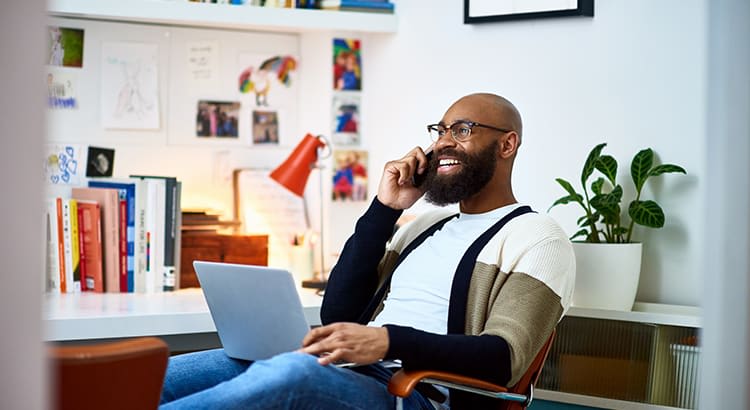 A person sitting at a desk, engaged in a conversation over the phone, appearing relaxed and productive. The setting seems to be an office or home workspace, with a laptop in front of them.